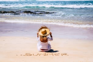Summer vacation holiday concept with people - white dress tourist woman sit down at the beach on the sand looking the blue waves from sea and holding the hat - summer write on the sand