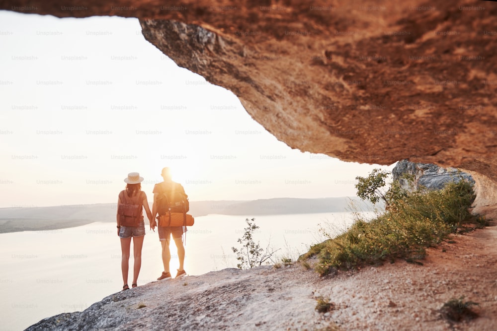 Dos turistas hombres y mujeres con mochilas se paran en la cima de la montaña y disfrutan del amanecer. Concepto de vacaciones de aventura de estilo de vida de viaje.