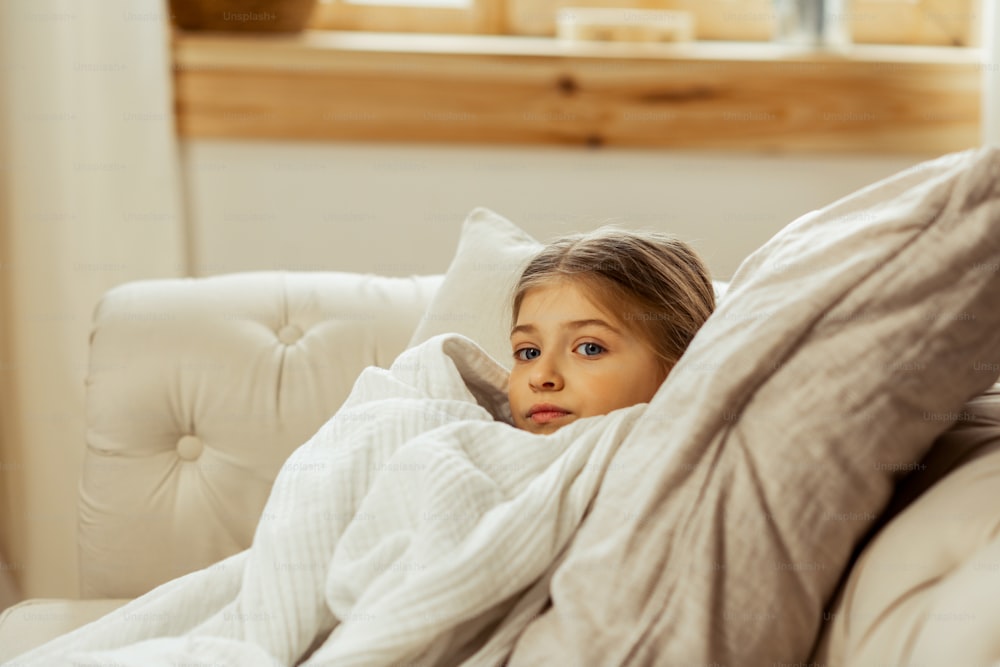 Uma menina doente na cama. Pequena menina doce e bonita de cabelos castanhos com um frio deitado na cama envolto em um cobertor quente