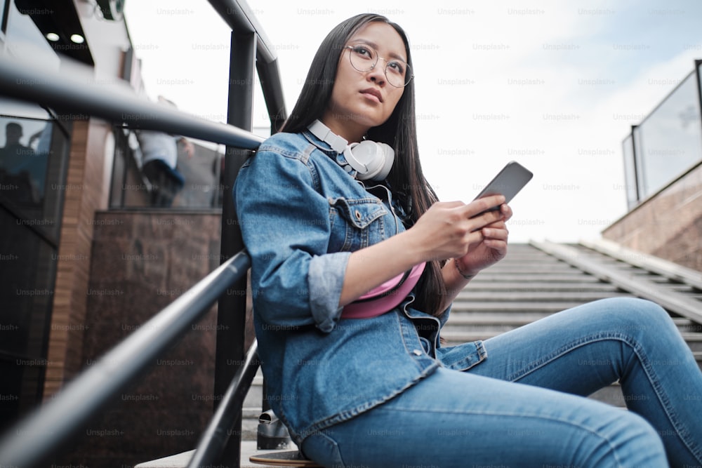 Close up of young asian skater girl sitting on her longboard on the stairs, using smart phone, wearing blue jeans, and pink belt bag, wearing round glasses and white headphones, looking away