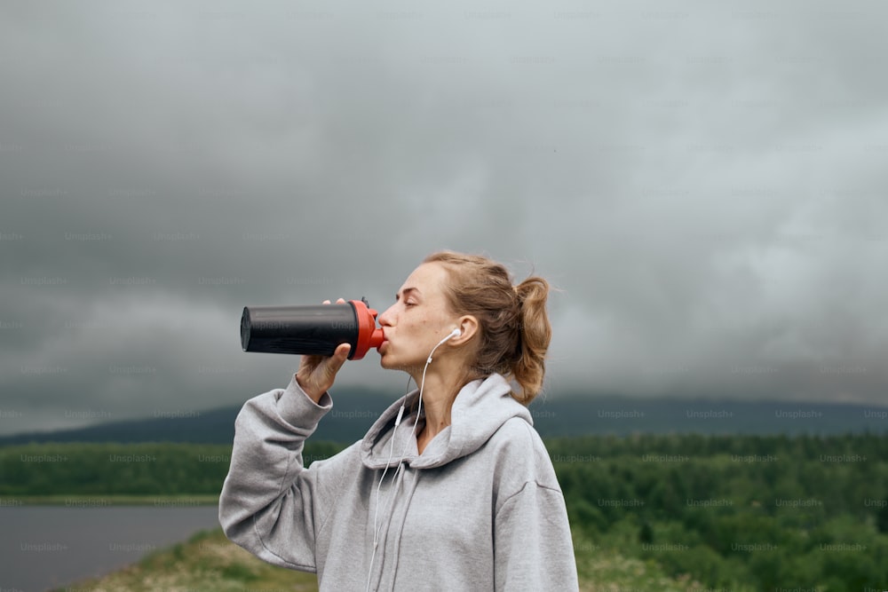 痩せた女性のスポーツウーマンは、暗い日に風光明媚な山の景色を見て、屋外のボトルから水を飲む