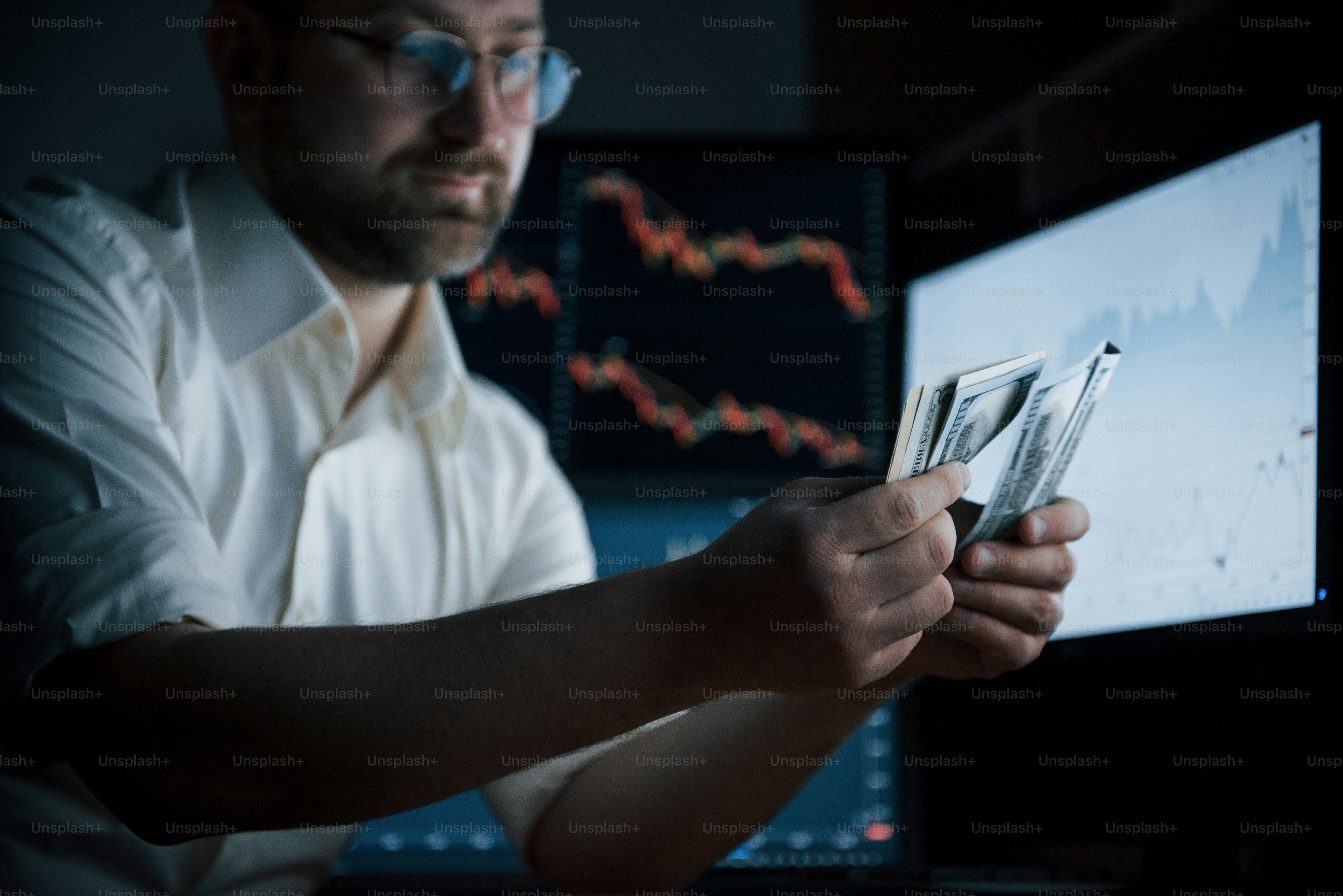 Hard earned money. Bearded man in white shirt works in the office with multiple computer screens in index charts.