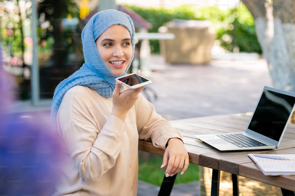 Chica feliz en hiyab grabando mensajes de voz en el teléfono inteligente mientras está sentada junto a la mesa frente a la computadora portátil en el parque