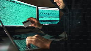 Rubare soldi. Vista laterale del giovane hacker barbuto in felpa nera con cappuccio che utilizza più computer e tiene in mano la carta di credito mentre è seduto in una stanza buia. Codice binario. Attacco informatico. Sicurezza informatica