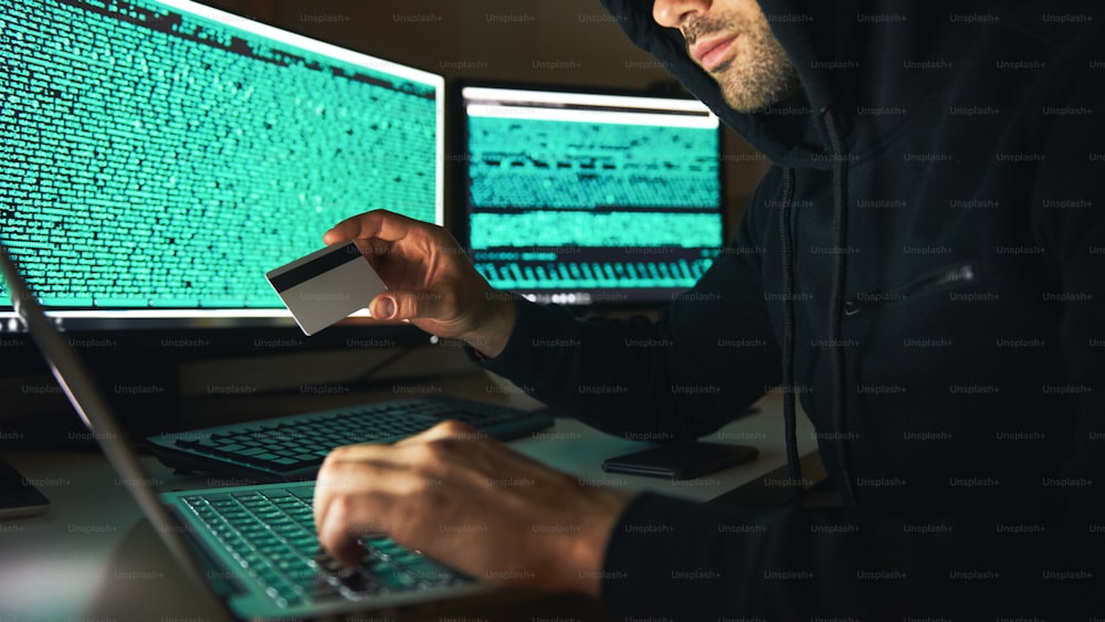 Rubare soldi. Vista laterale del giovane hacker barbuto in felpa con cappuccio nera che utilizza più computer e tiene in mano la carta di credito mentre è seduto in una stanza buia. Codice binario. Attacco informatico. Sicurezza informatica