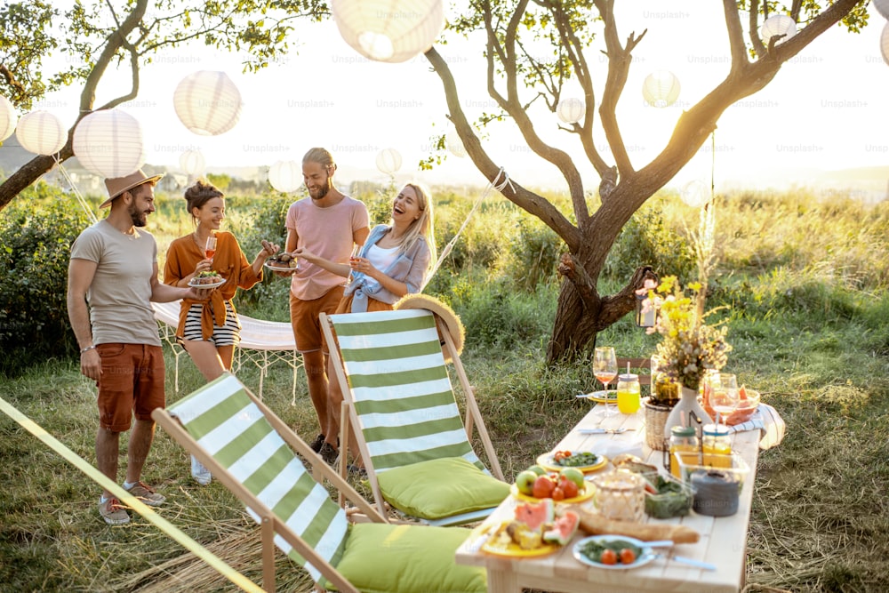Gruppe junger und glücklicher Freunde, die Spaß bei einem festlichen Picknick im wunderschön dekorierten Garten mit Mittagstisch bei Sonnenuntergang haben