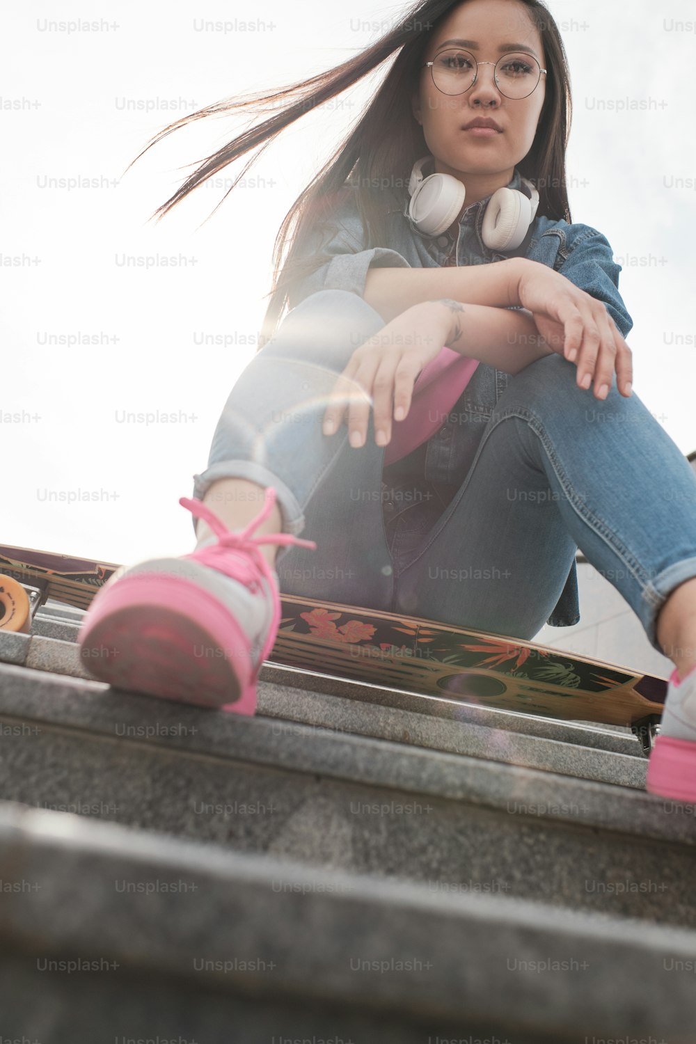 Junges Mädchen von unten fotografiert, wie sie draußen mit einem Longboard auf der Treppe sitzt.