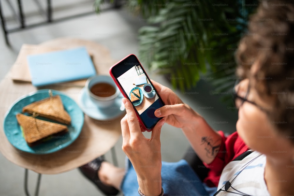 Mujer móvil que usa su teléfono inteligente para tomar fotos de sándwiches y beber en la mesa mientras está sentada en un café