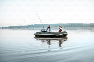 Grandfather with adult son fishing on the inflatable boat on the lake with calm water early in the morning. Wide landscape view