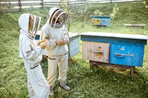 Two young beekepers in protective uniform working on a small apiary farm with wooden beehives on the background