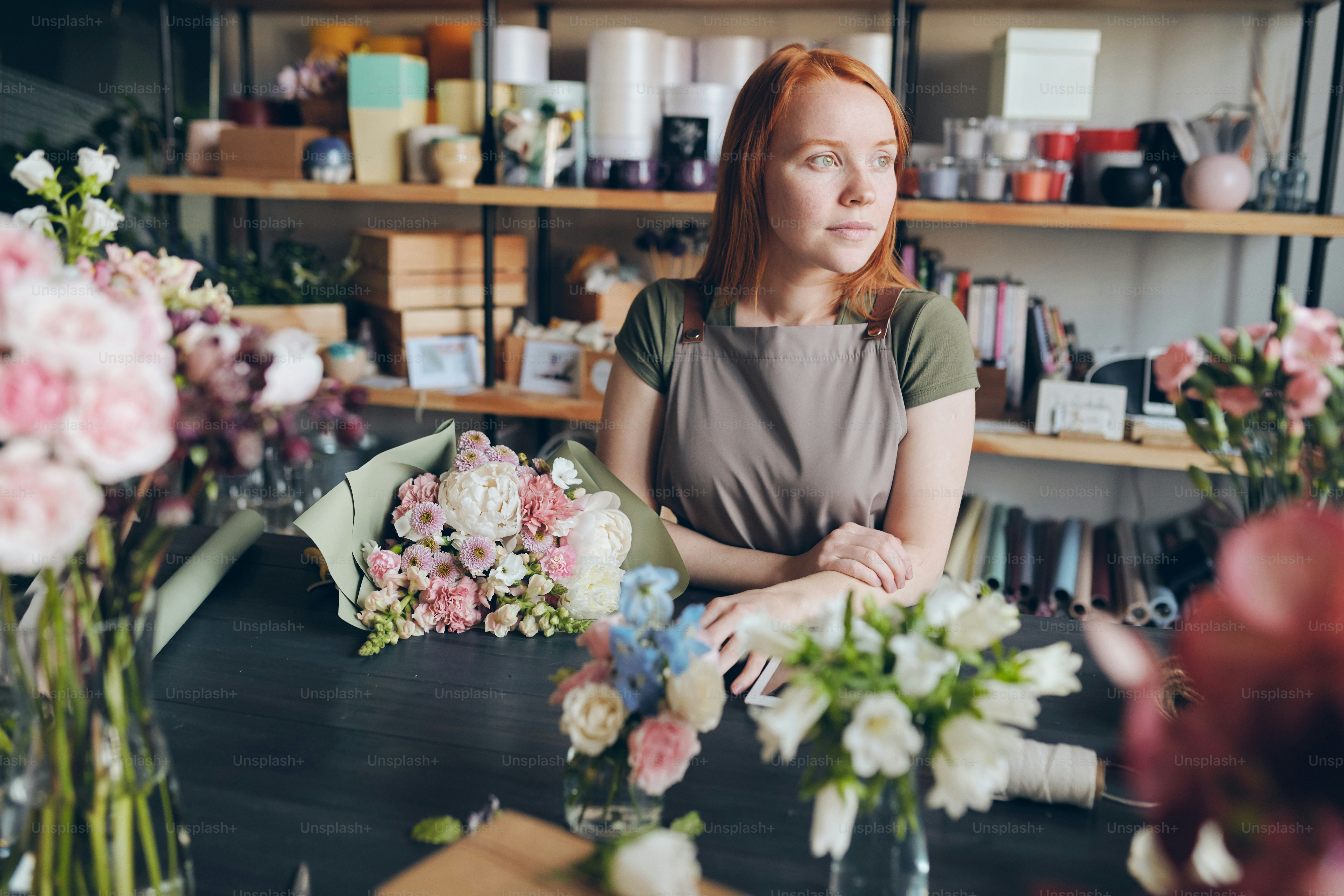 Pensive beautiful florist in apron leaning on counter with tender bouquet and waiting for customer in flower shop