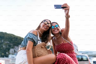 Happy young female friends in sunglasses sitting and taking a selfie on mobile phone on blurred background of city street