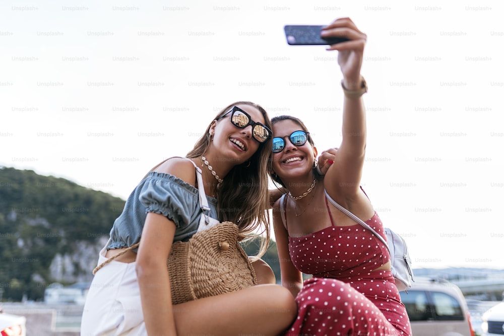 Happy young female friends in sunglasses sitting and taking a selfie on mobile phone on blurred background of city street