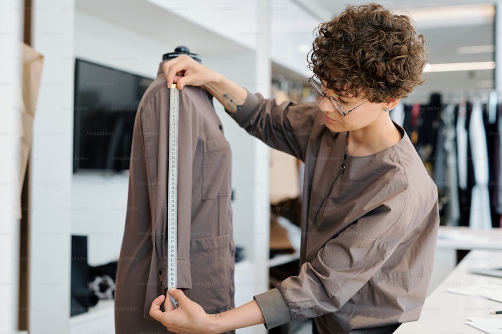 Jeune femme brune bouclée mesurant la longueur des manches de la veste sur mannequin tout en travaillant dans son studio