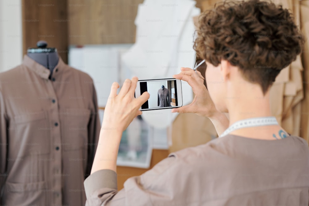 Young woman making shot of unfinished dress or jacket on mannequin to send it to one of clients