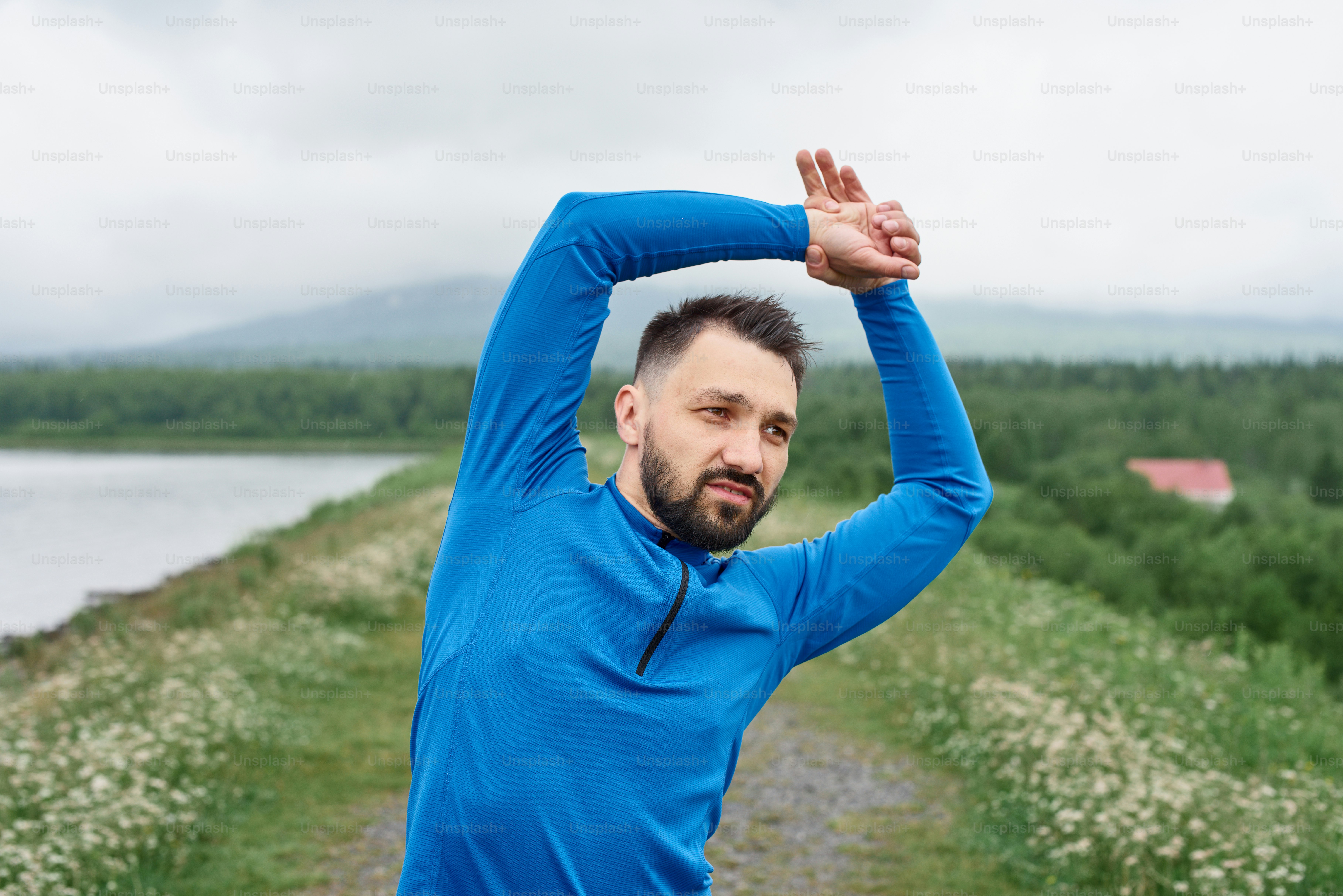 屋外で運動し、腕の筋肉をウォームアップし、夏に、憂鬱な日に、風光明媚な景色を望む道路に立ち、ウォームアップストレッチを行い、青いジャケットを着て、あごひげを生やしている中年のスポーツマン