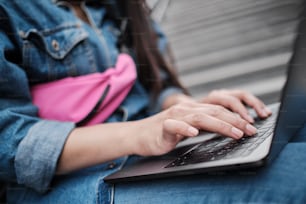 Close up of female freelancer working on laptop outdoors, typing on keyboard, she's wearing blue denim and pink belt bag