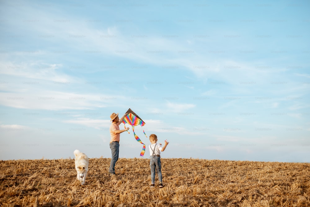 Pai e filho lançando pipa de ar colorida no campo, cachorro passeando por aí. Conceito de uma família feliz se divertindo durante a atividade de verão