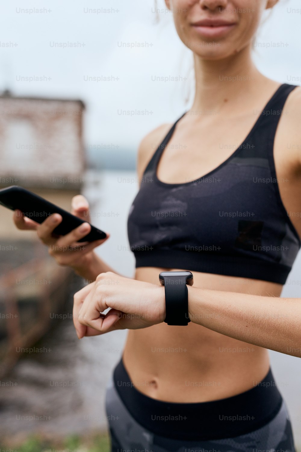 Portrait of mid age skinny sports woman checking smart watch with smart phone, synchronizing, exercising outdoor in summer, on gloomy day with scenic view, wearing tank