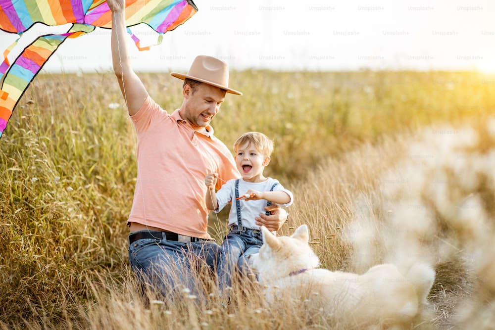 Retrato de um pai e filho felizes segurando pipa de ar colorida enquanto sentado junto com o cão no campo durante o pôr do sol