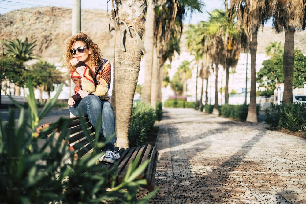 alone and sitting woman in a park with a lot of palms on the background with thoughts and she's thinking about something or looking at the street - casual lifestyle outdoor leisure actiity