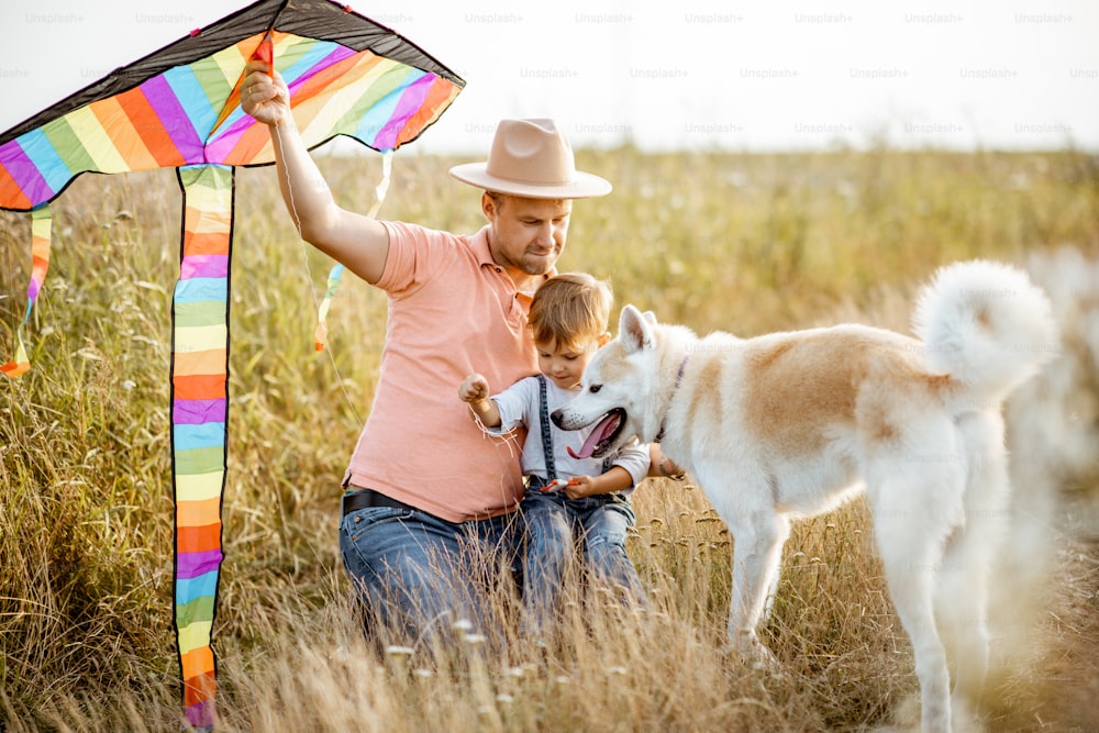 Portrait of a happy father and son holding colorful air kite while sitting together with dog on the field during the sunset