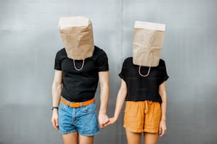 Portrait of a young couple with paper bags on their heads, keeping hands together on the grey wall background