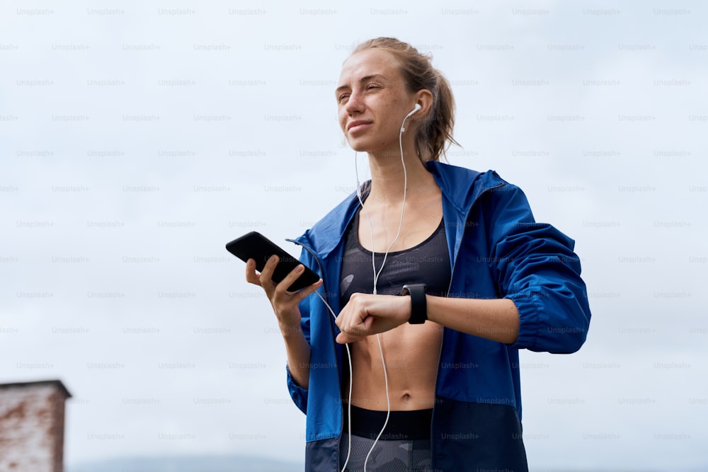 Portrait of mid age skinny sports woman with earphones synchronizing smart watch with smart phone outdoor in summer, on gloomy day with scenic view, wearing tank and blue rain coat
