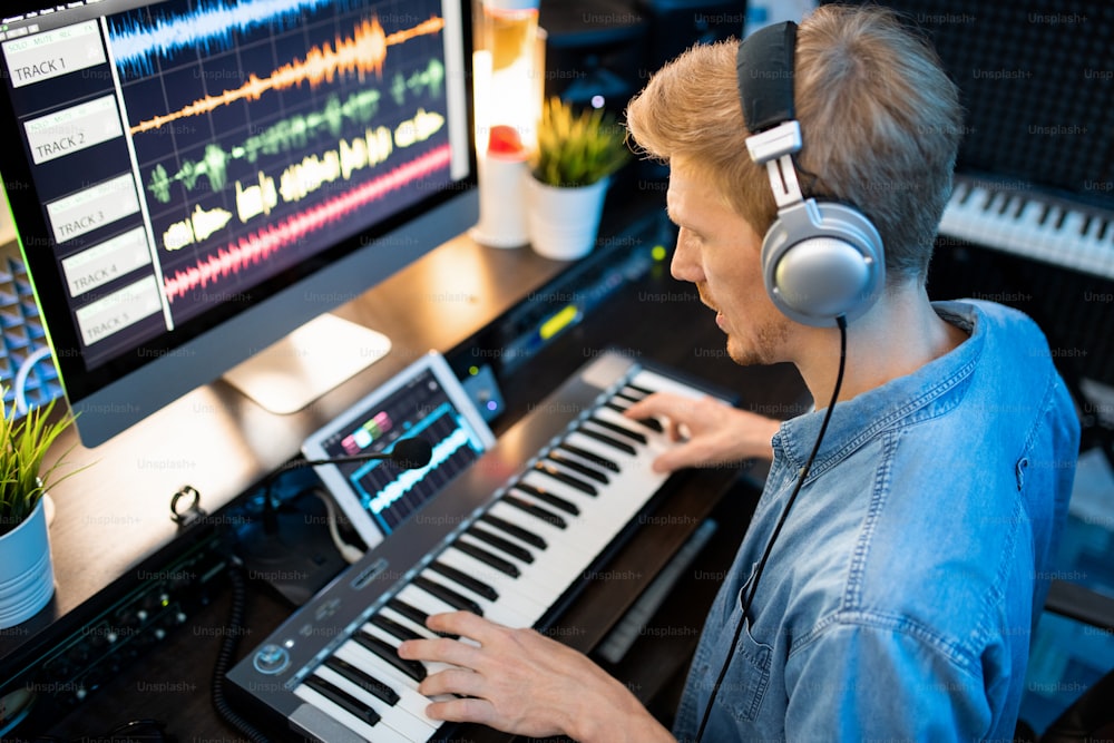 Joven músico contemporáneo en auriculares grabando nueva música y canciones mientras toca en el teclado del piano en el estudio