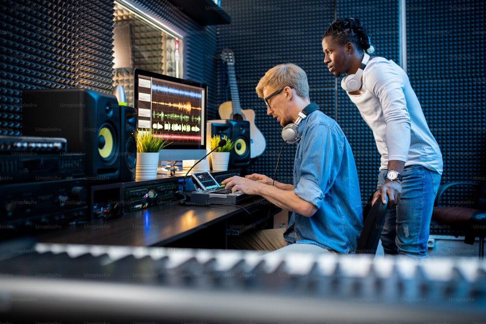 One of young intercultural musicians pressing pianoboard keys while his colleague looking at computer screen during teamwork in studio