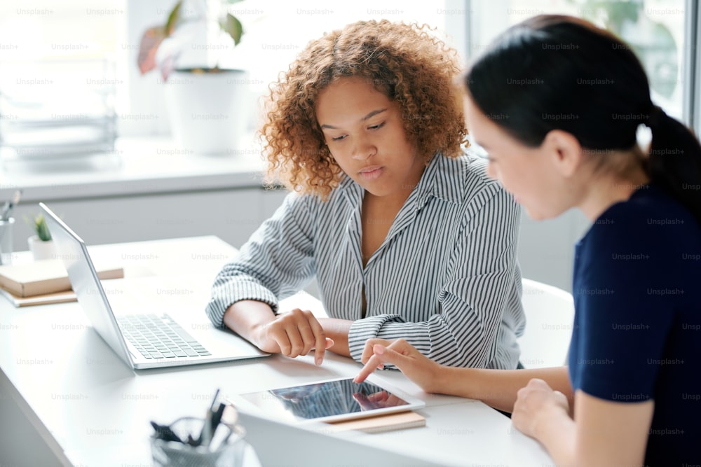 Young confident businesswoman pointing at tablet screen while showing her trainee or subordinate how to work with data