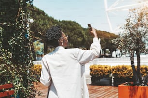 Rearview of an elegant black girl shooting during a sightseeing on a camera of her smartphone; a fancy young African female in a white trench and sunglasses is photographing cityscape via a cellphone