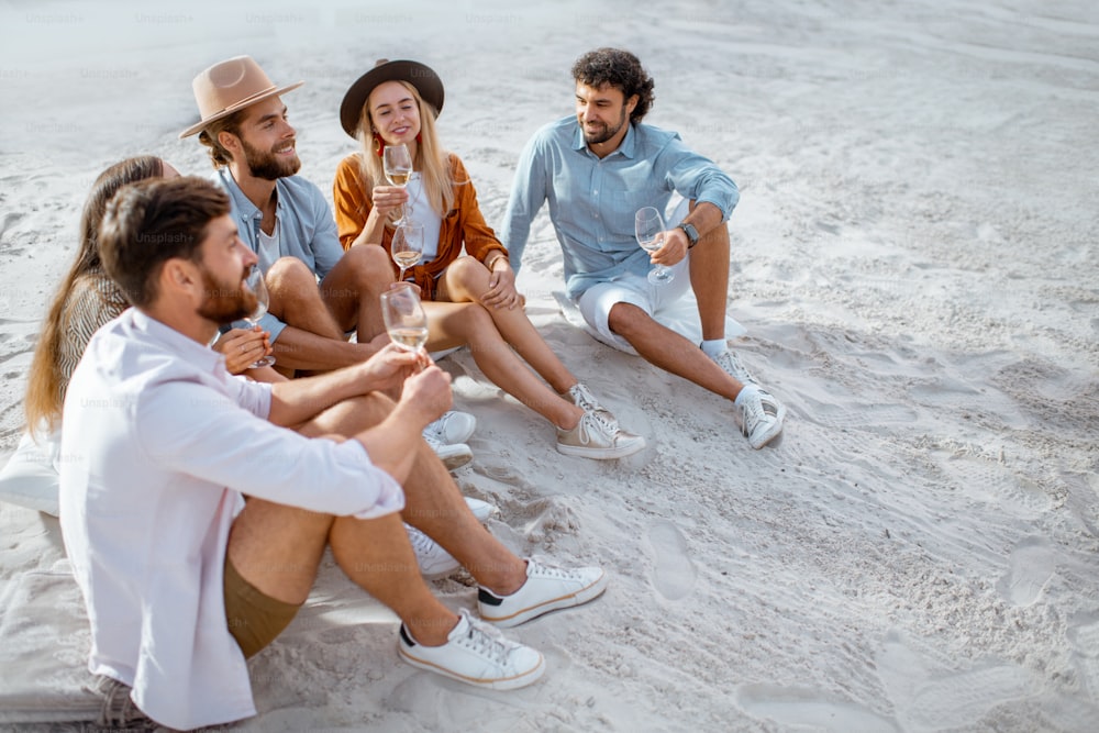 Group of young friends hanging out together with wine glasses, having a festive meeting and enjoying evening time on the sandy beach