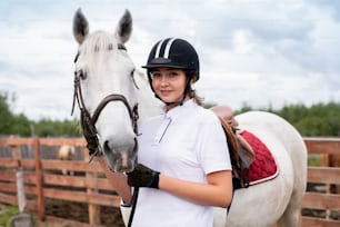 Mulher sorridente nova na roupa equestre olhando para você enquanto está perto do cavalo de corrida branco na frente da câmera