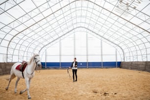 Jeune femme active avec des brides debout sur une arène sablonneuse et regardant un cheval de course blanc de race pure chevauchant autour