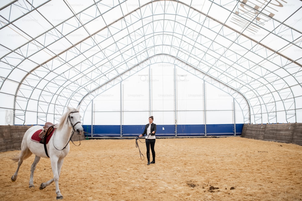 Aktive junge Frau mit Zaumzeug steht auf sandiger Arena und schaut auf weiße, reinrassige Rennpferde, die herumreiten