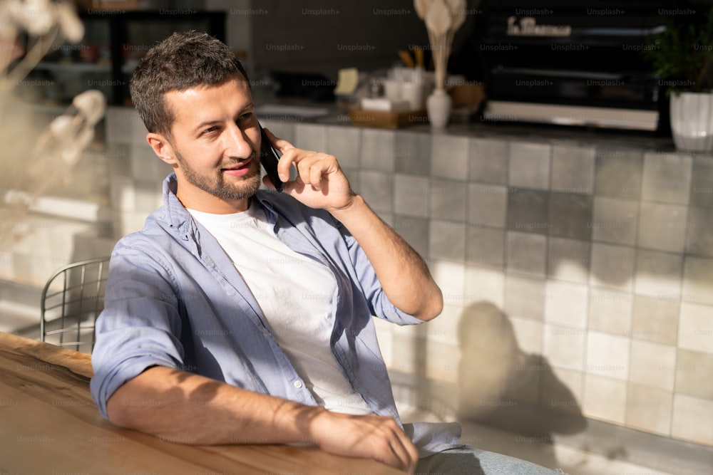 Happy young businessman with smartphone talking to colleague or consulting client while relaxing by table in cafe