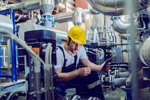 Focused plant worker in overalls, with protective helmet on head and antiphons on ears using tablet for checking machine while crouching.