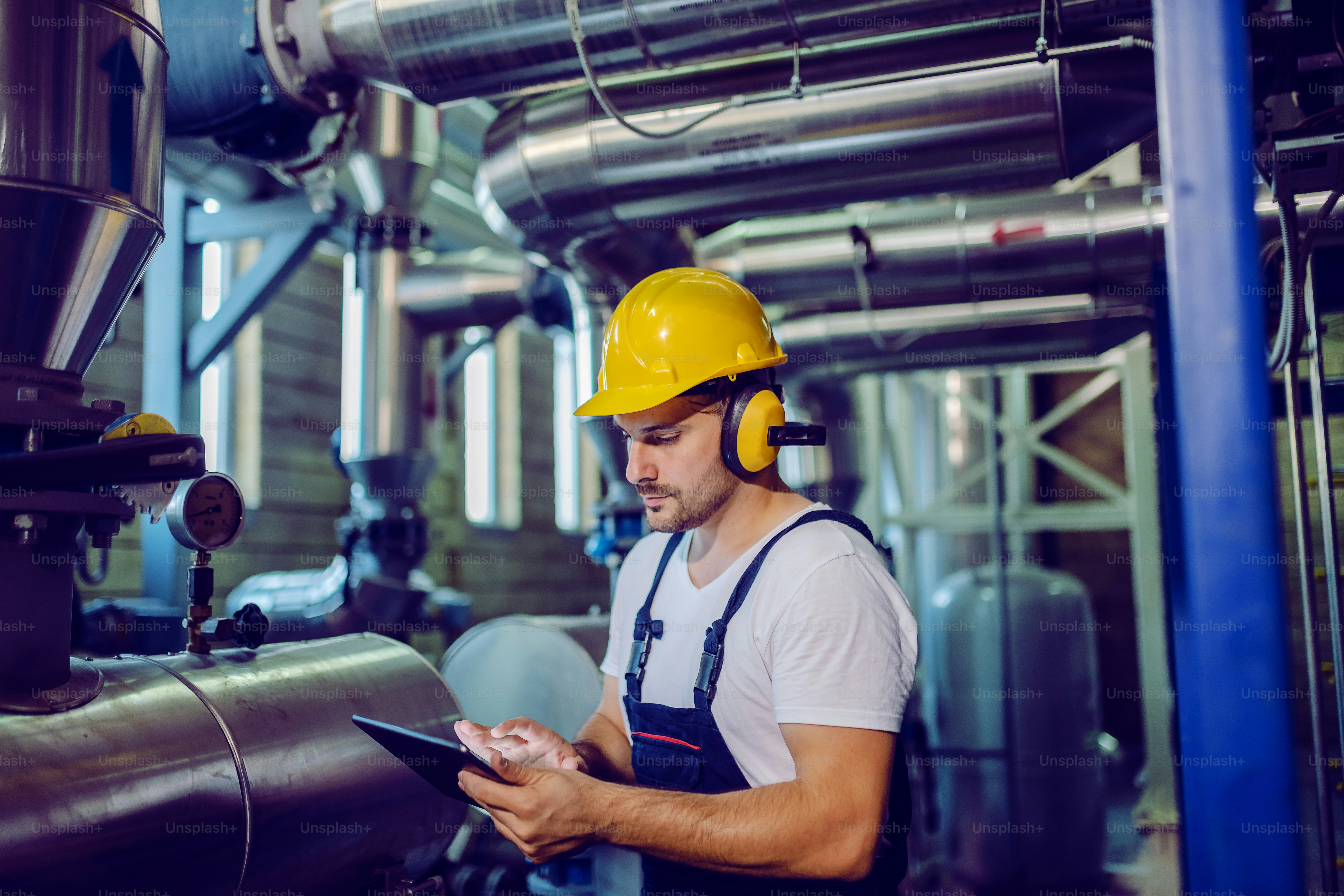Focused plant worker in overalls, with protective helmet on head and antiphons on ears using tablet for checking machine.