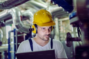 Focused plant worker in overalls, with protective helmet on head and antiphons on ears using tablet for checking machine.