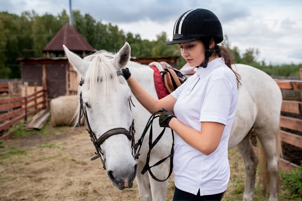 Menina ativa no capacete equestre e camisa polo branca e seu cavalo de corrida movendo-se para baixo do campo no ambiente rural