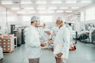 Dos supervisores hablando sobre la calidad de la comida. El más joven sostiene la tableta mientras que el mayor sostiene la carpeta con gráficos. Ambos están vestidos con uniformes estériles y con redecillas para el cabello. Interior de la planta alimenticia.