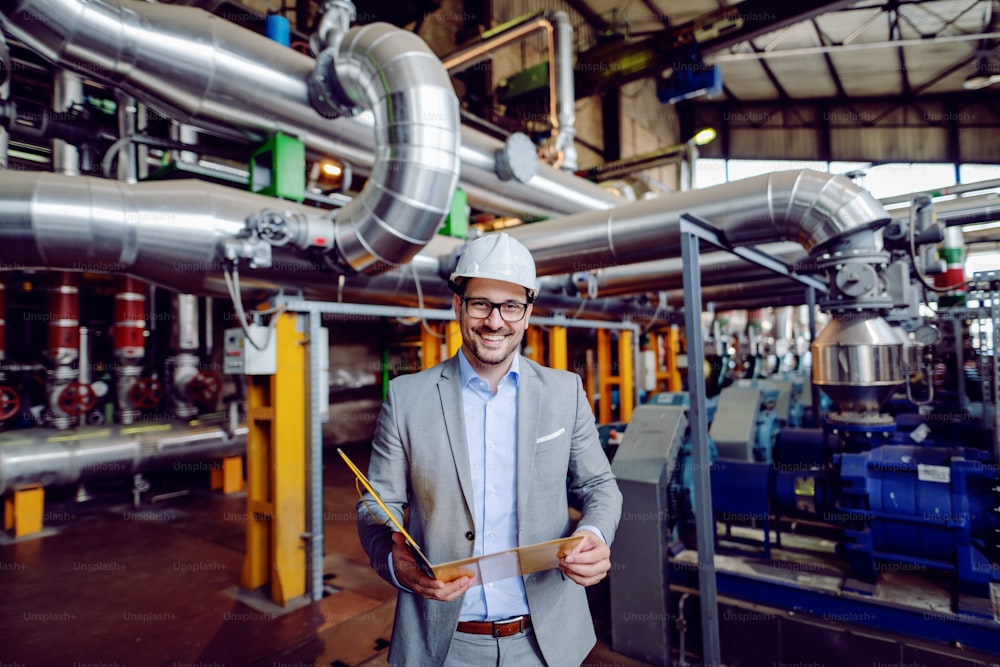 Handsome caucasian businessman in suit and with protective helmet on head holding folder with documents and looking at camera while standing in power plant.