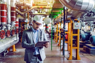 Handsome caucasian manager in gray suit and with helmet on head using tablet while standing in power plant.