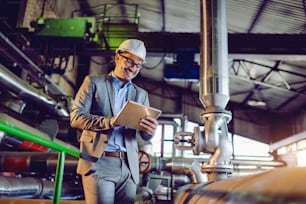 Handsome smiling caucasian supervisor in suit and with helmet on head using tablet while standing in power plant.