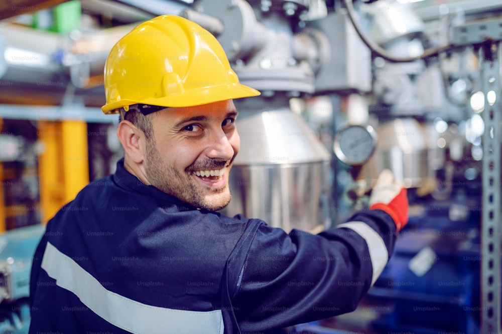 Smiling hardworking energy plant worker in working suit screwing valve while looking at camera.