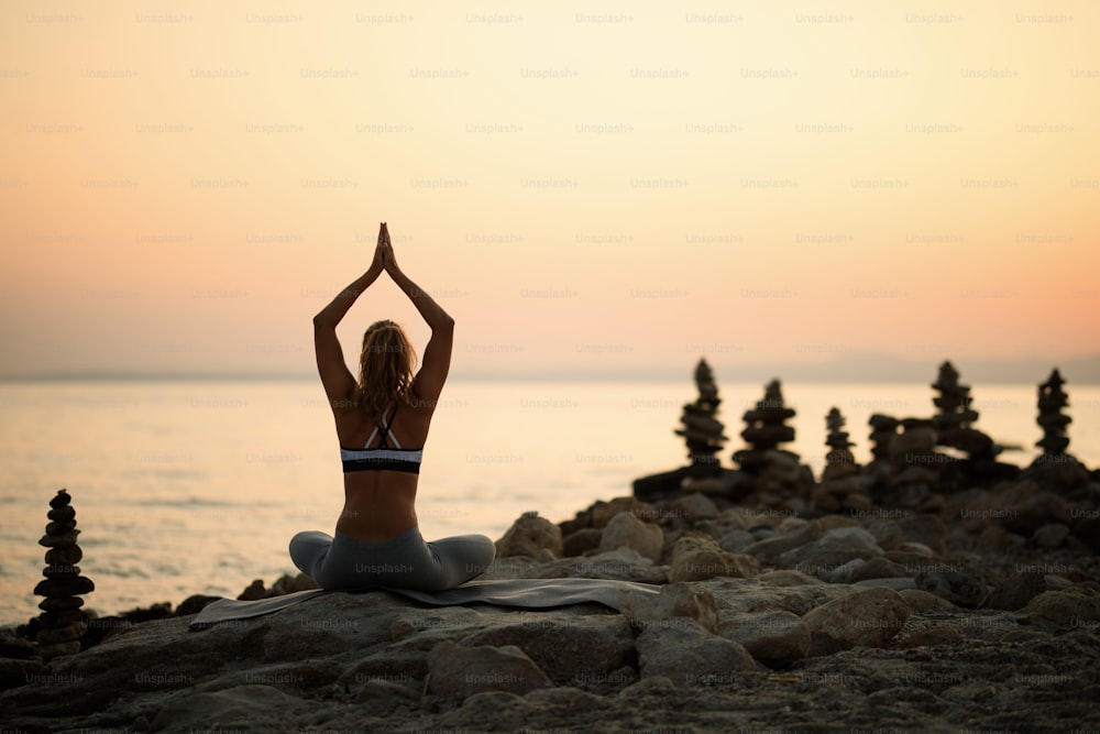 Back view of woman with hands above head meditating on a rocky beach at sunset. Copy space.