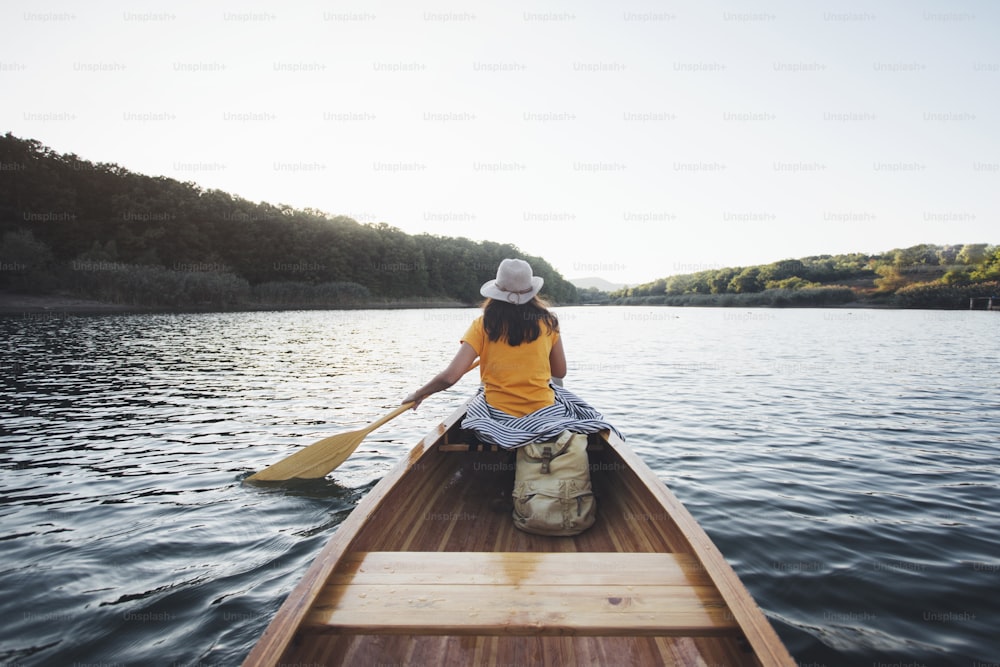 Vista trasera de una chica hipster remando en la canoa en el lago de la puesta de sol.
