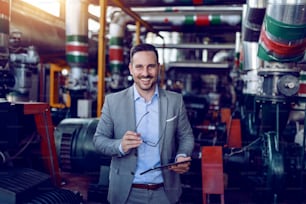 Supervisor caucasiano sorridente em terno cinza segurando tablet e óculos nas mãos e olhando para a câmera enquanto está em pé na usina de energia.