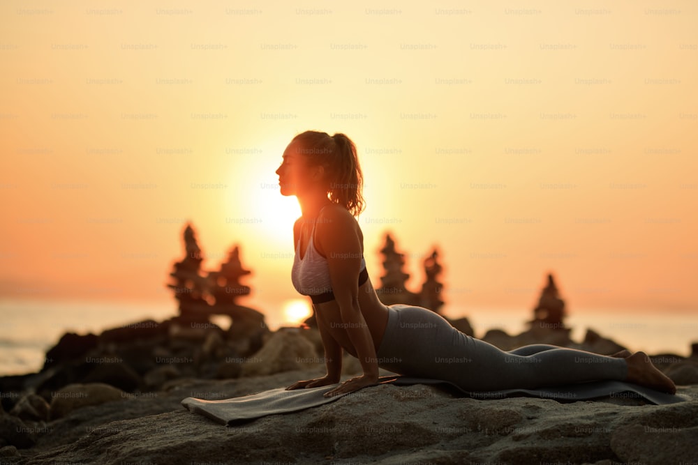 Young athletic woman doing relaxation exercises and stretching on rocky beach at sunset. Copy space.